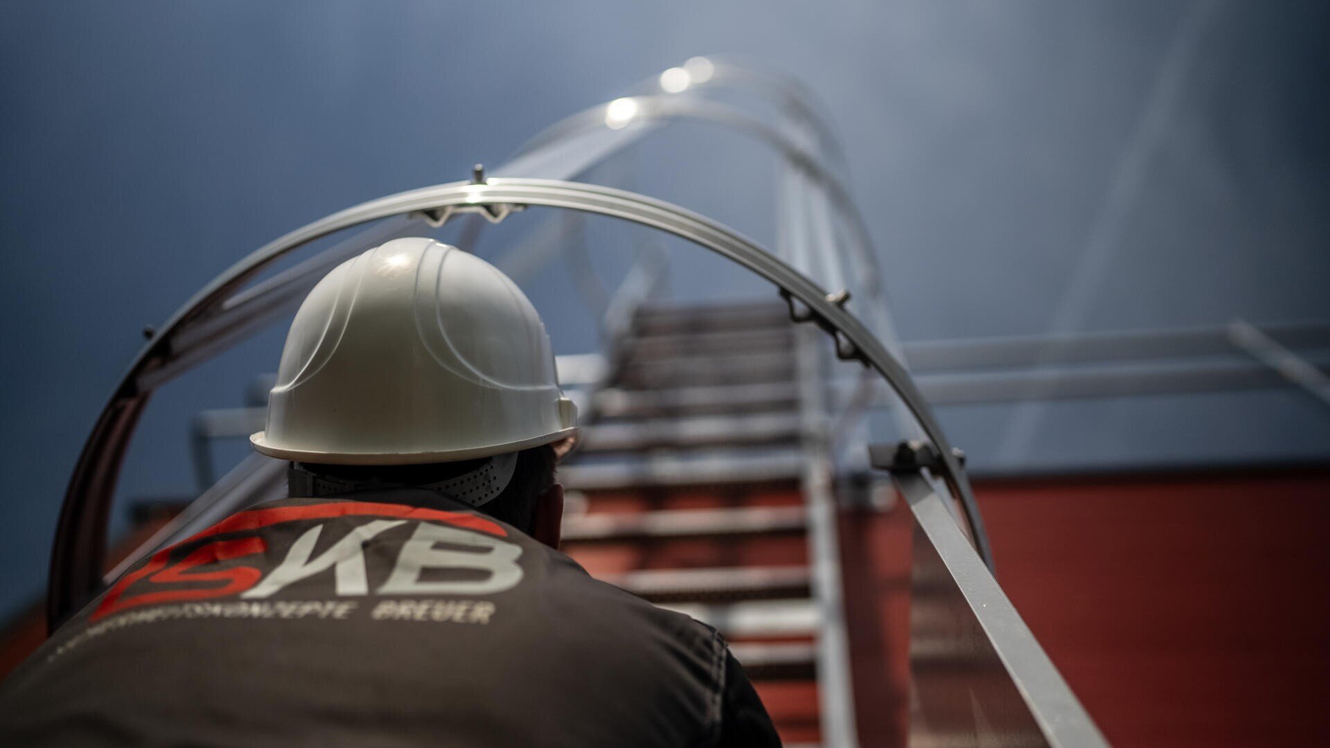 Eine Person, die einen weißen Schutzhelm und dunkle Arbeitskleidung trägt, steht am Fuß einer Metallleiter, die an einem Gebäude befestigt ist, und blickt gegen einen bewölkten Himmel nach oben.