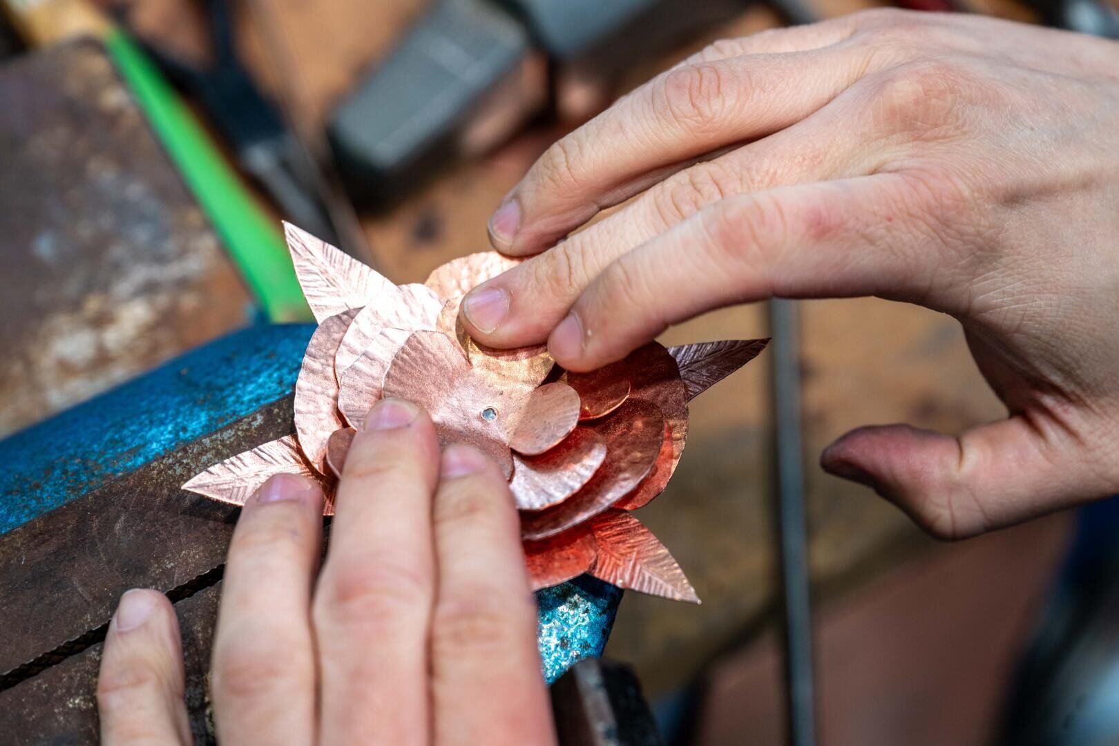 Die Hände einer Person montieren oder justieren geschichtete, kupferfarbene Metallblütenblätter in Form einer Blume, die in einem blauen Schraubstock auf einer Werkbank gehalten werden. Werkzeuge und andere Materialien sind im unscharfen Hintergrund zu sehen.