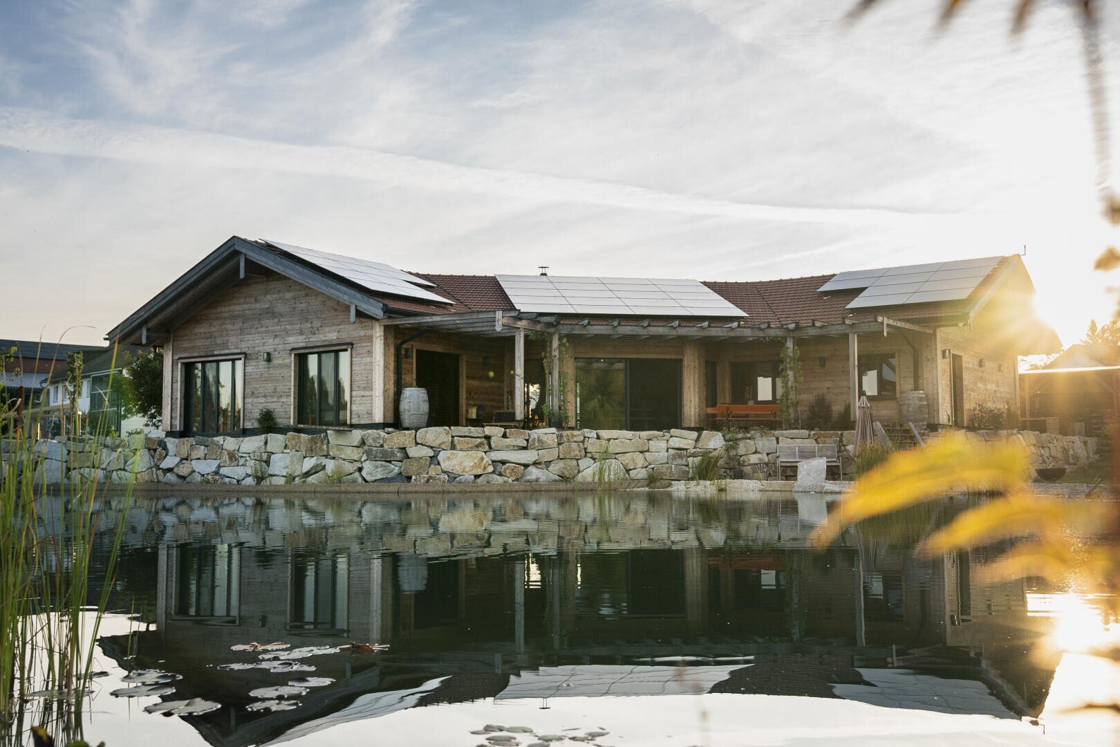 A modern house with solar panels on the roof sits next to a tranquil pond that reflects the building and the clear sky at sunset. The exterior is characterized by stone walls, large windows and a natural landscape.