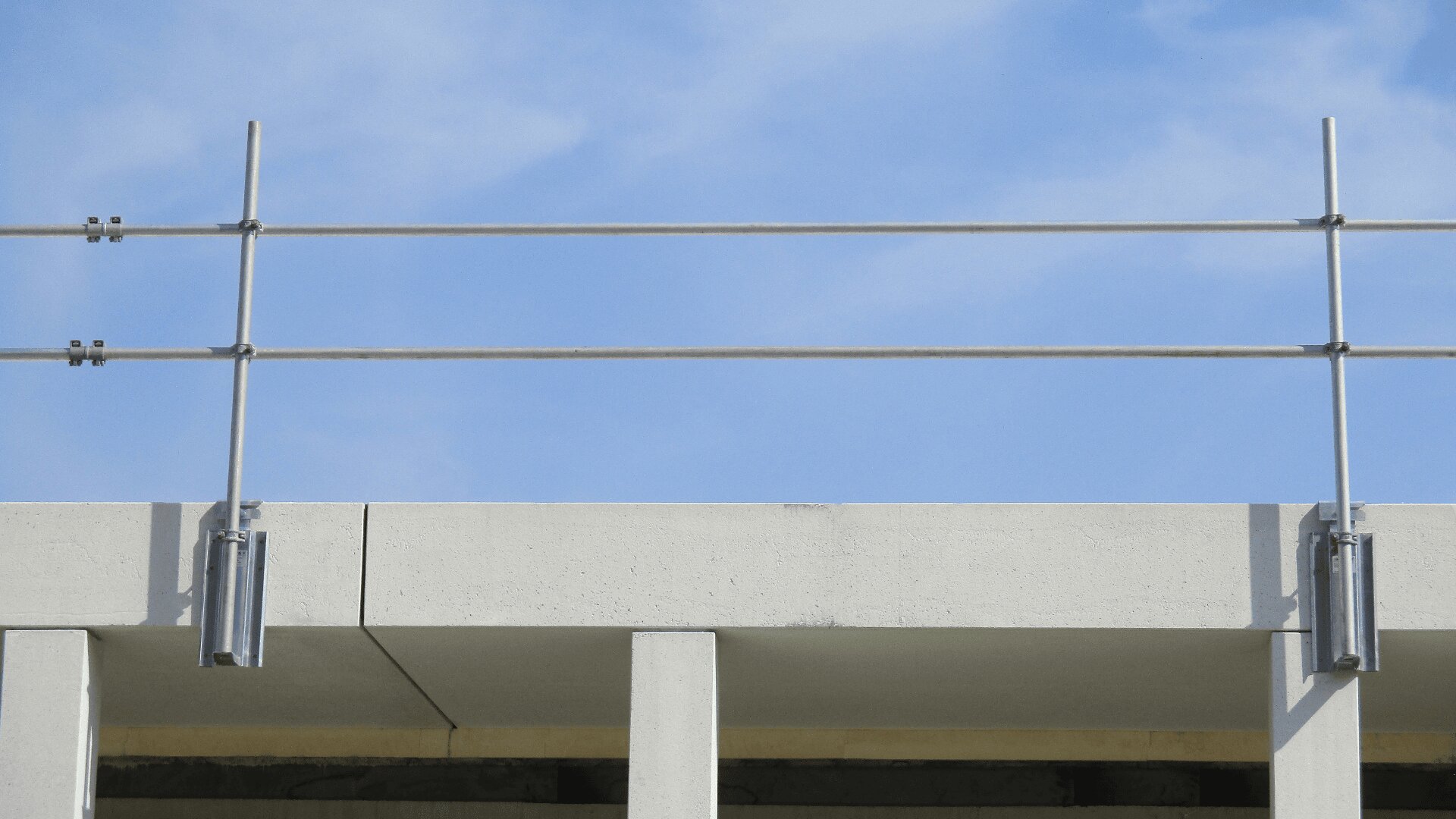 Eine Nahaufnahme eines im Bau befindlichen weißen Betongebäudes mit einem Sicherheitsgeländer auf dem Dach vor blauem Himmel. Die Struktur hat vertikale und horizontale Betonbalken.