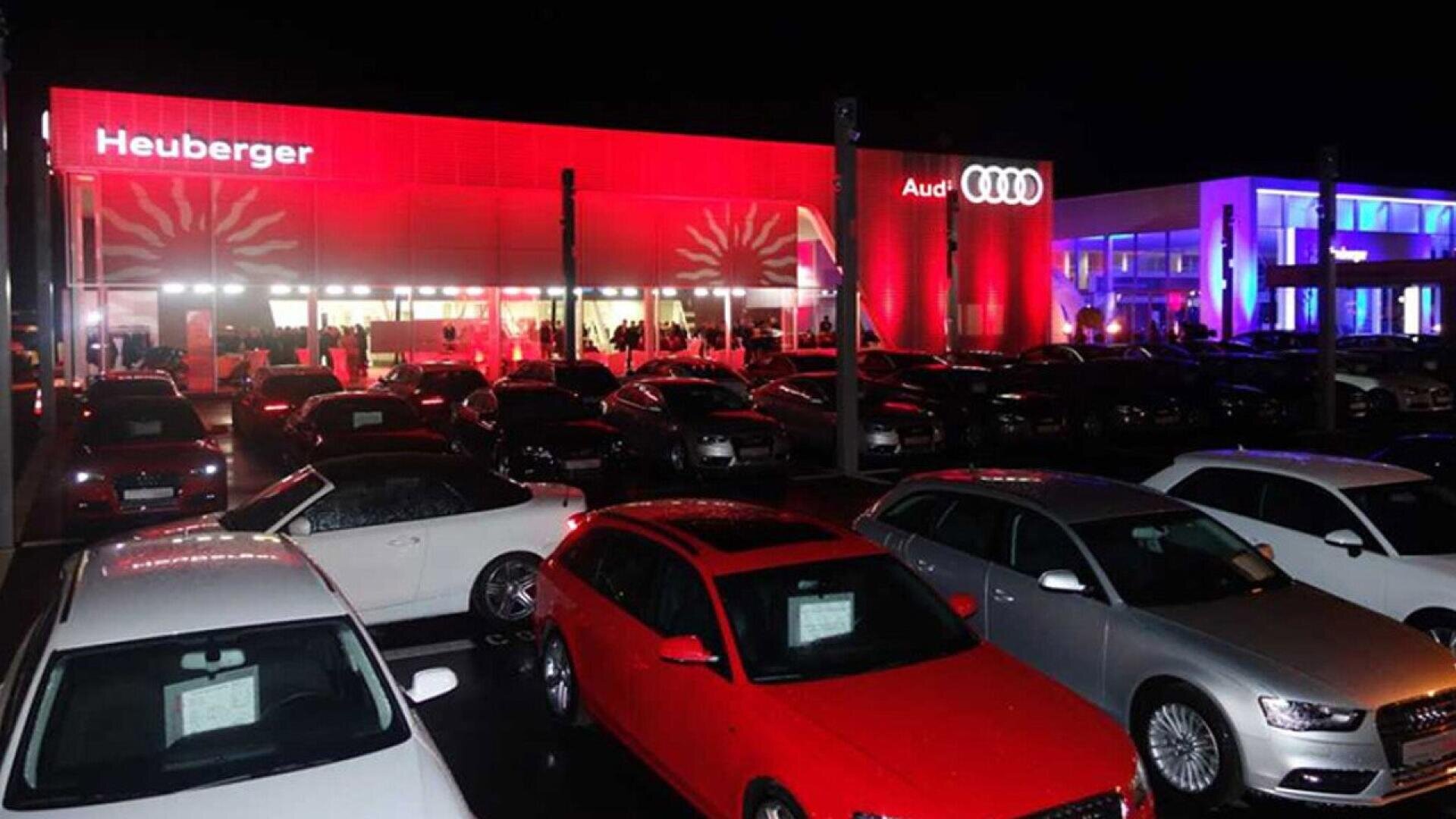 A brightly lit Audi dealership at night, with the building illuminated in red and blue lights. Several cars are parked in rows in the foreground, and people are visible inside the building.