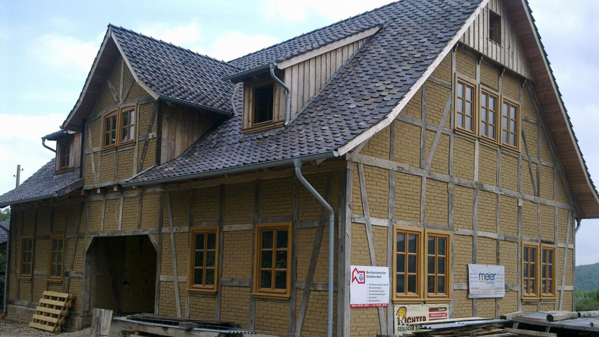 A timber-framed house with a dark tiled roof and multiple windows, some with shutters. The house is under renovation, with construction signs and materials visible in front. The sky is partly cloudy.