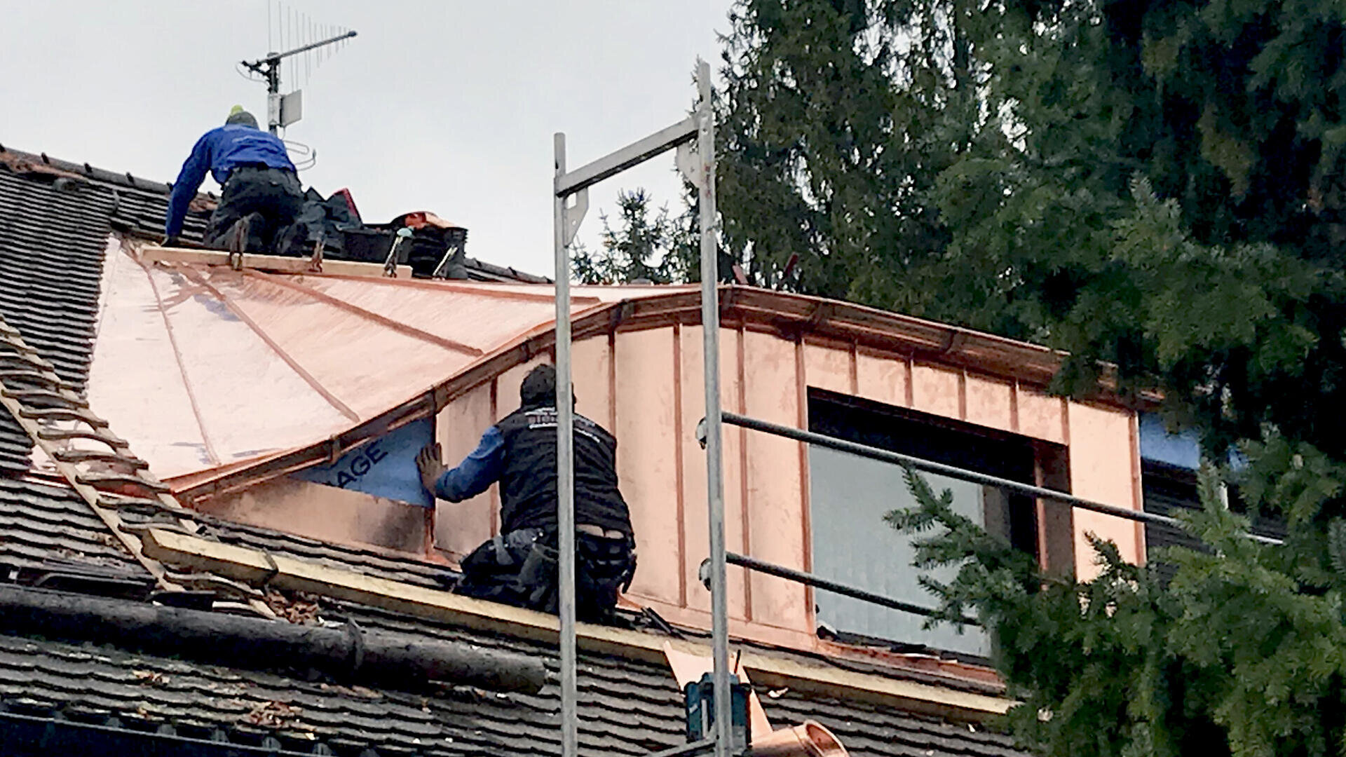 Two workers on scaffolding and a rooftop install copper sheets on a house dormer. One worker is kneeling on the roof, while the other works near a window. Trees and construction materials surround the area.
