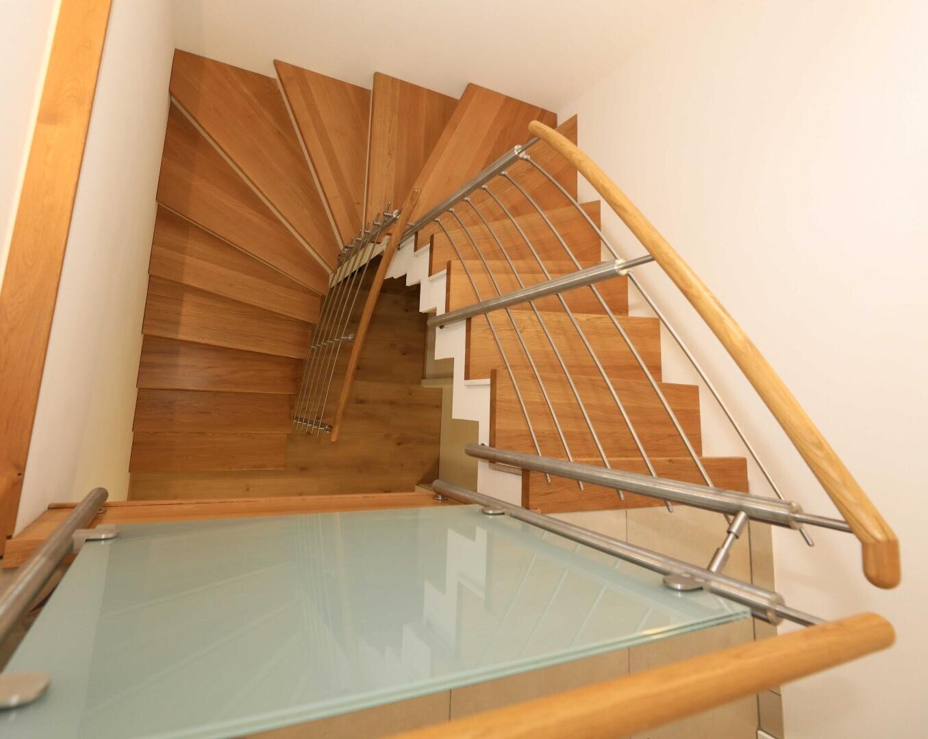 Draufsicht auf eine moderne Wendeltreppe aus Holz mit Metall- und Holzgeländer vor hellen Wänden. Der Treppenabsatz hat eine Glas- oder Milchglasoberfläche.