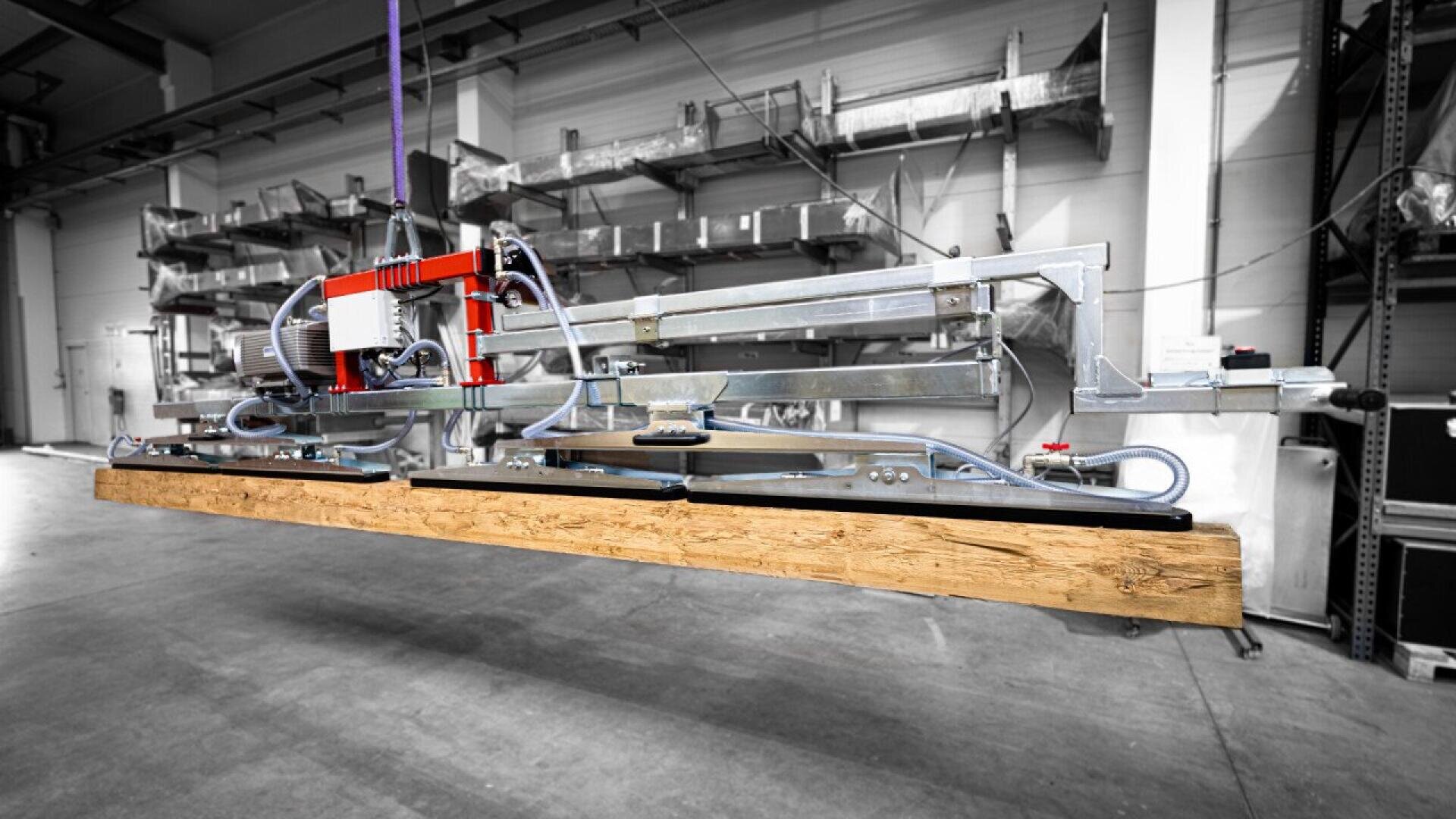 A large industrial machine with metal parts and wiring is lifting a long wooden beam inside a warehouse with shelves in the background.