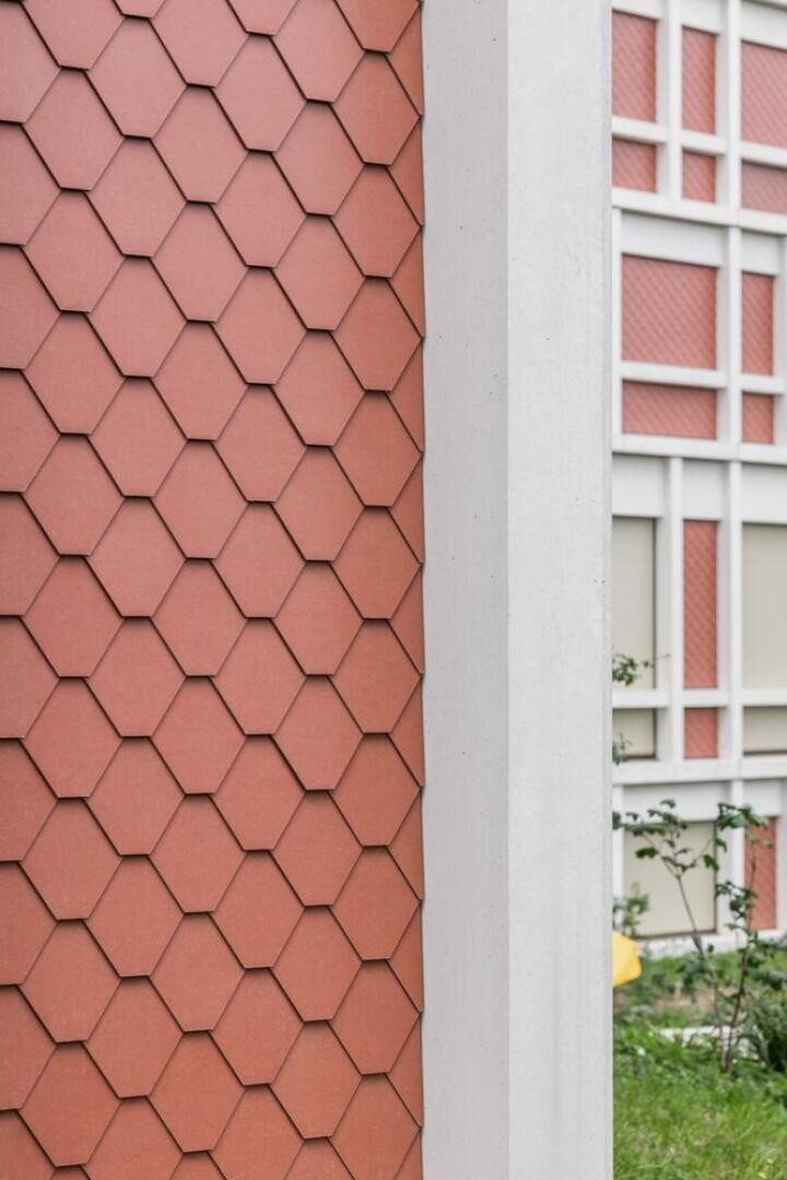 Close-up of a building façade with red hexagonal tiles on the left and a white concrete column in the middle, with a wall with a grid pattern and some grasses and plants in the background.