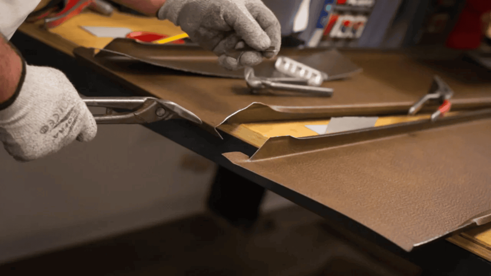A person wearing gray gloves uses metal snips to cut a sheet of brown metal, while another hand holds a small piece over a workbench with tools, including a hammer and a metal straight edge.
