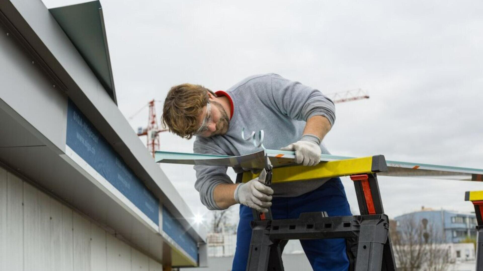 A man wearing safety glasses and gloves uses a tool to work on a metal sheet, standing on a ladder near the edge of a building under construction, with cranes visible in the background.