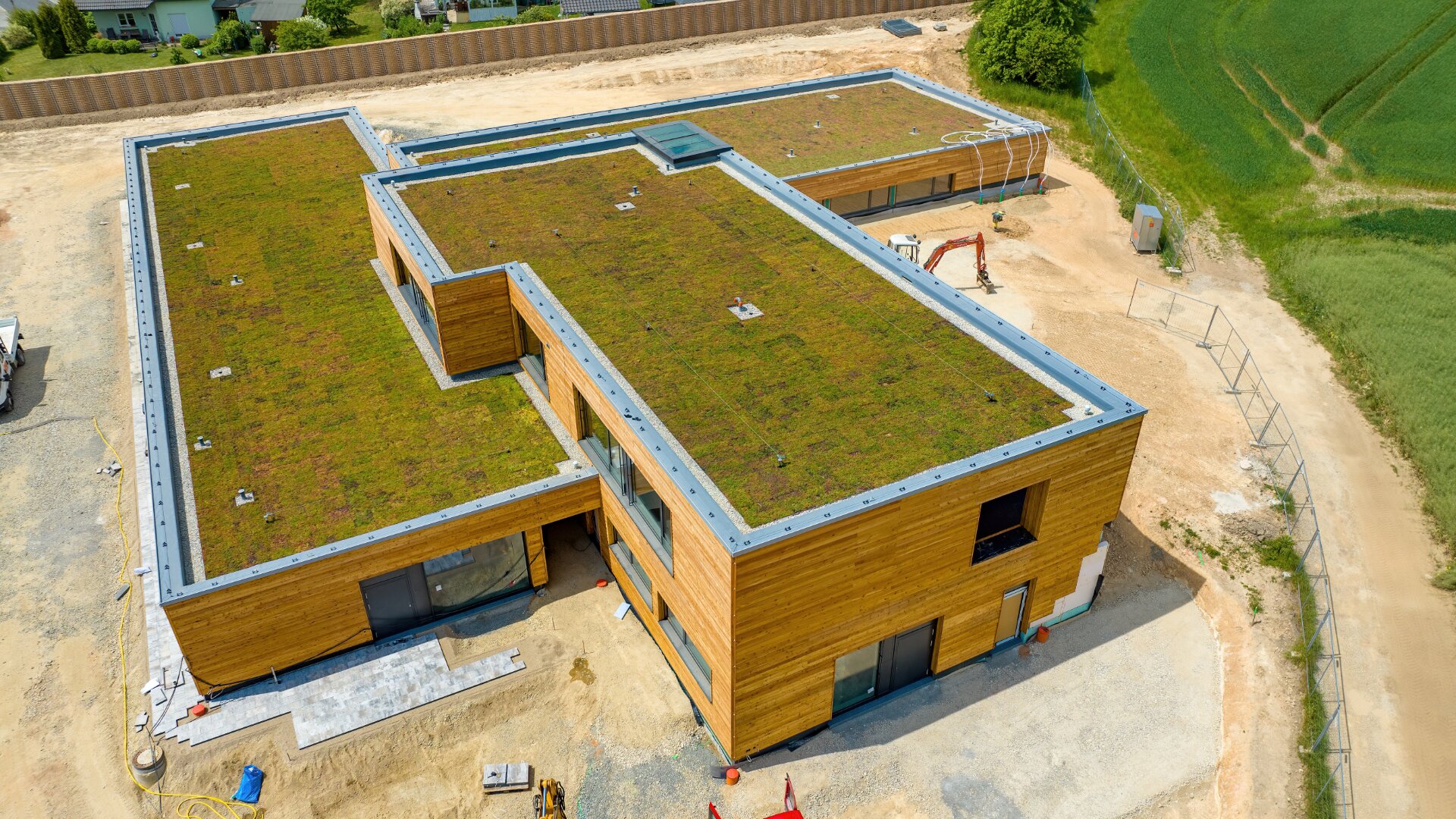 Luftaufnahme eines modernen L-förmigen Gebäudes mit Gründach, Holzfassade und großen Fenstern, umgeben von Erde und einigen Baumaschinen, neben Grasfeldern.
