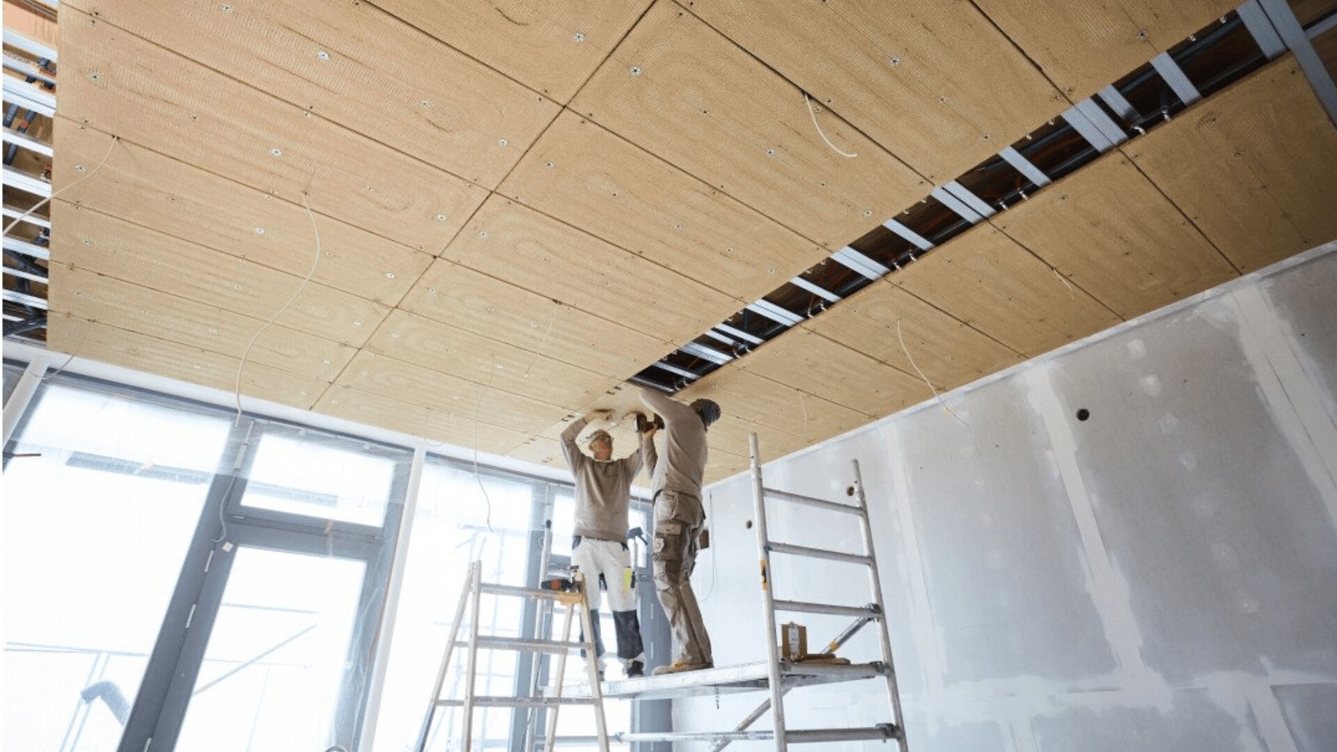 Two construction workers stand on ladders, installing large ceiling panels in a room under renovation. The room has unfinished walls, exposed wiring, and construction materials visible.