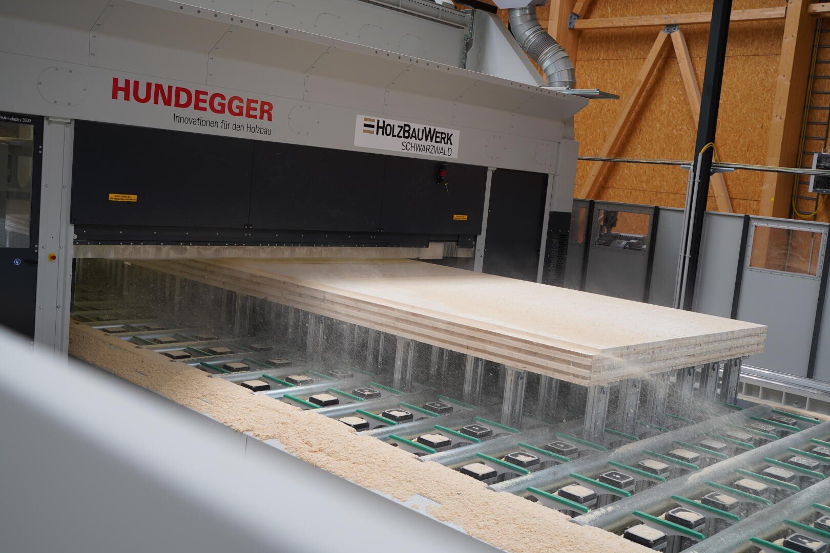 Eine Hundegger-Holzbearbeitungsmaschine bearbeitet große Holzplatten in einem modernen Sägewerk. Auf dem darunterliegenden Förderband sind Holzspäne sichtbar. Die Szene spielt im Innenbereich, im Hintergrund sind freiliegende Holzwände zu sehen.