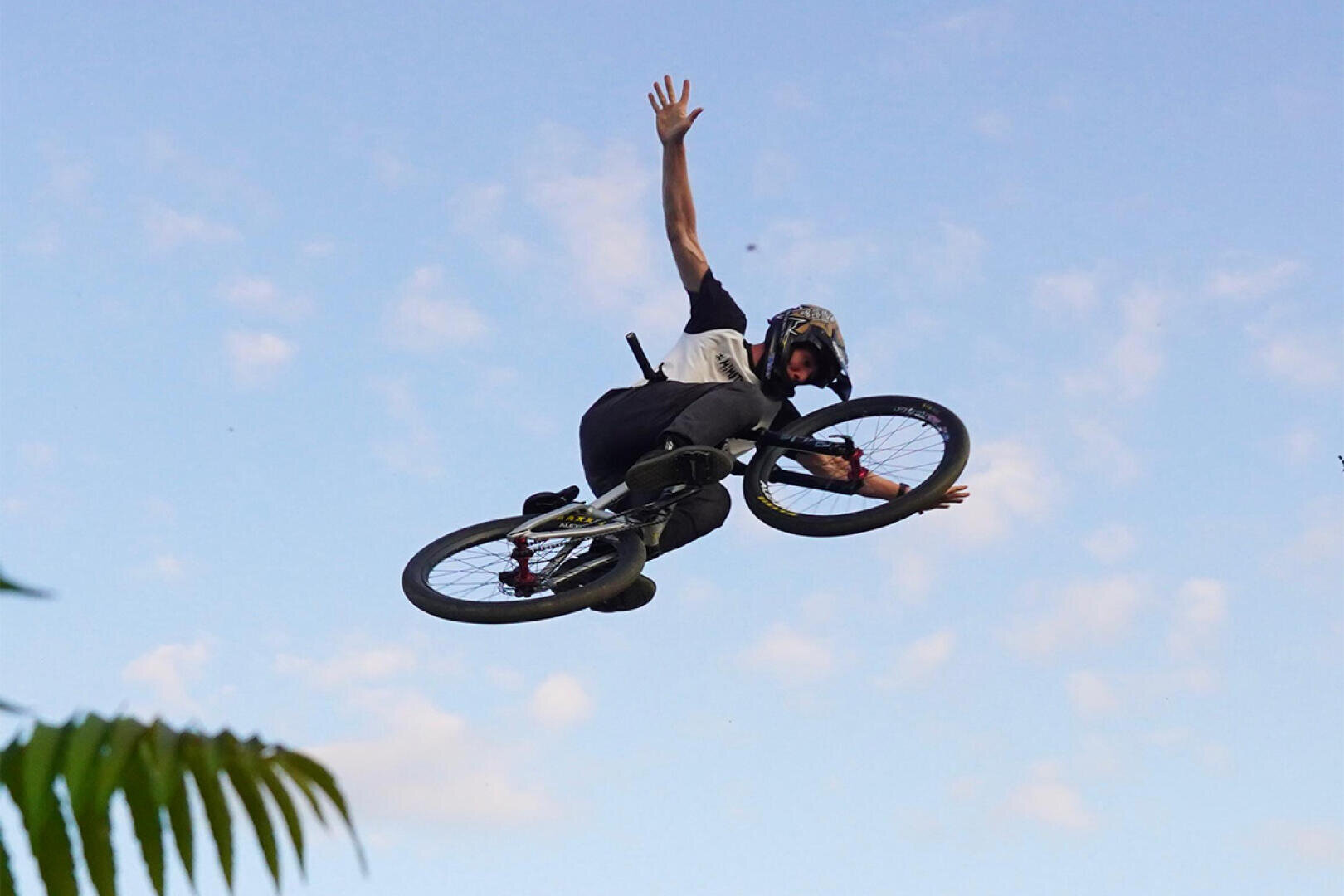 Eine Person, die einen Helm trägt, führt einen Trick auf einem BMX-Rad aus, in der Luft mit einem ausgestreckten Arm vor einem blauen Himmel mit vereinzelten Wolken. In der Ecke des Bildes sind grüne Blätter zu sehen.