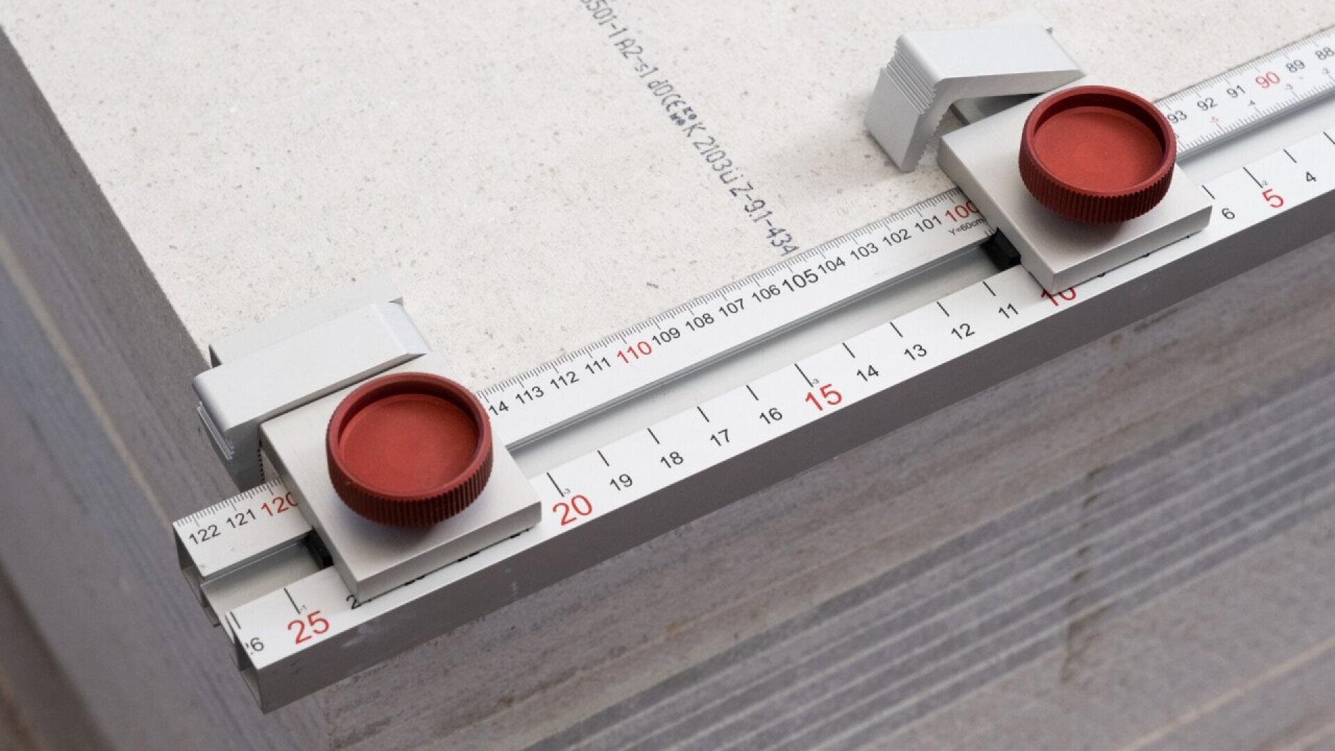 Nahaufnahme eines Messwerkzeugs mit Schiebeskala, roten Einstellknöpfen und voreingestelltem Lineal, das an der Kante gestapelter Baumaterialplatten positioniert ist. Das Werkzeug zeigt die Maße in Zentimetern an.