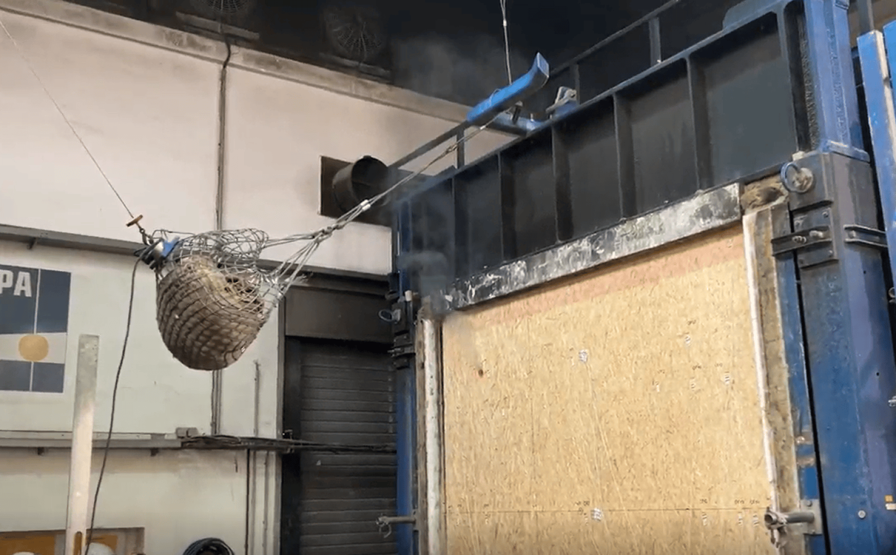 A large metal basket filled with objects hangs from cables in an industrial warehouse near a high, partially open wooden door, from which steam or smoke can be seen rising in the background.