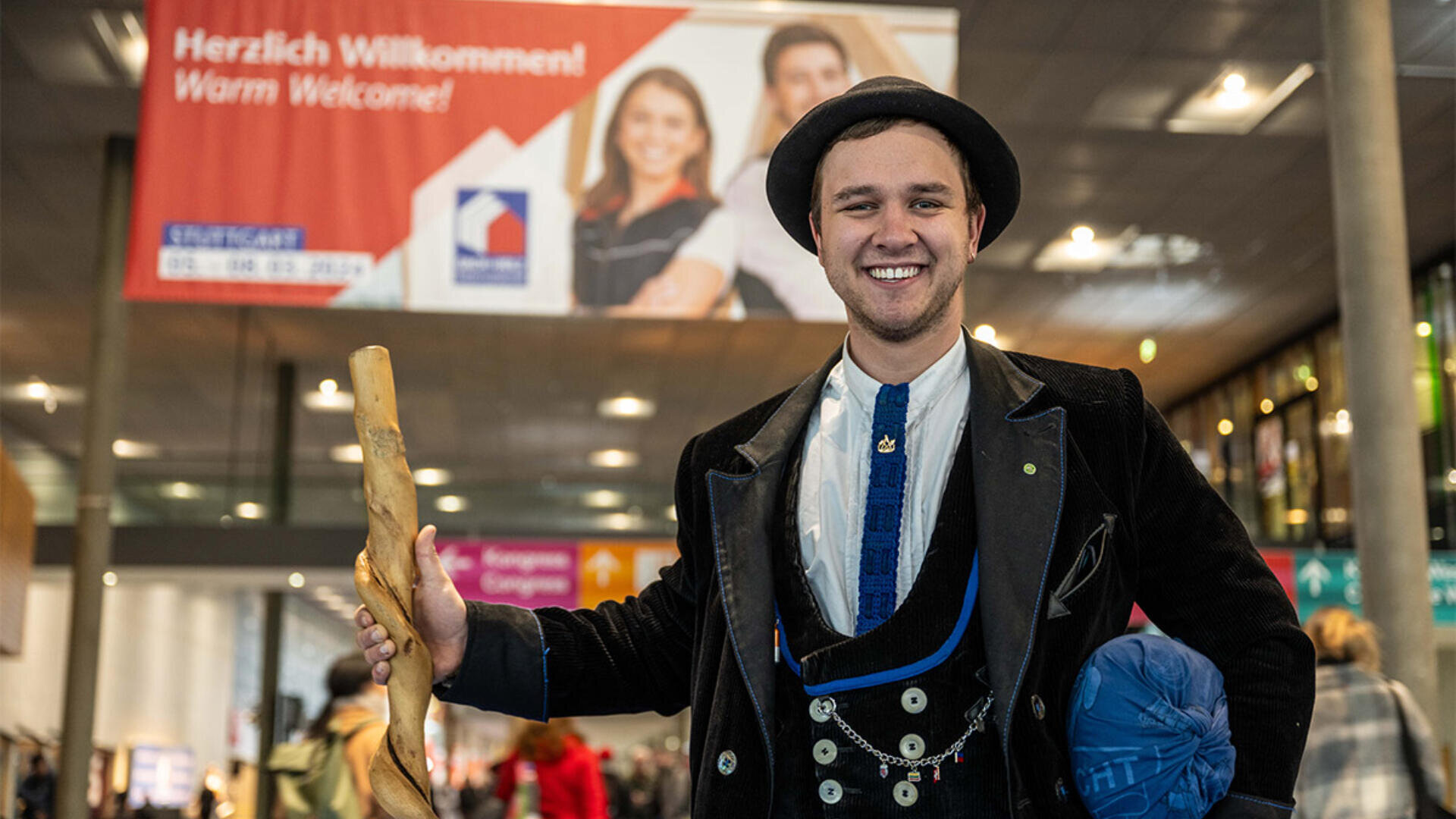 Ein lächelnder junger Mann in traditioneller deutscher Kleidung hält einen Holzstab in einem öffentlichen Innenraum, im Hintergrund sind ein großes Willkommensbanner mit der Aufschrift „Herzlich Willkommen!“ und Menschen zu sehen.