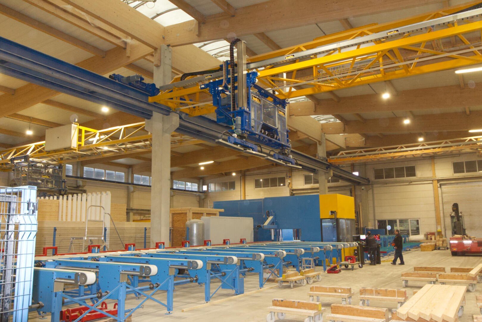 Ein modernes Fabrikinterieur mit automatisierten Maschinen, Förderbändern und Stapeln von Holzbrettern unter hellen Deckenleuchten, in denen Arbeiter die Maschinen bedienen.