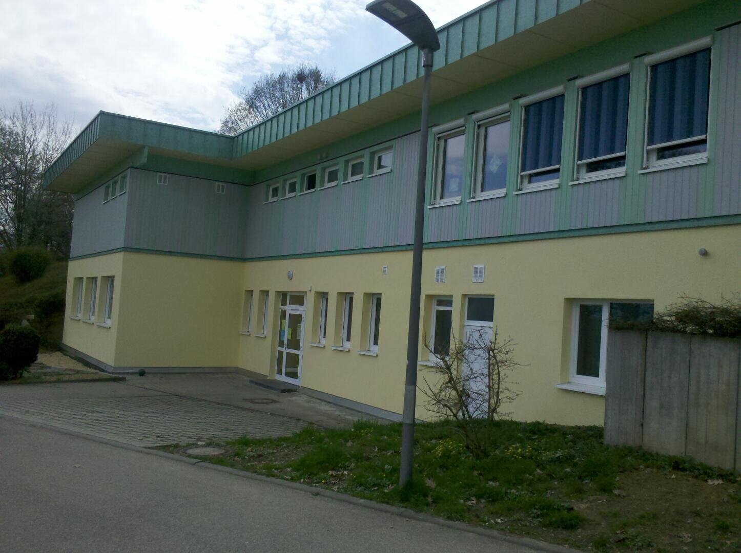 Ein zweistöckiges modernes Gebäude mit hellgelben Wänden im Erdgeschoss und hellgrünen Akzenten darüber, mit vielen Fenstern, einer Glaseingangstür und einer Straßenlaterne davor an einem bewölkten Tag.