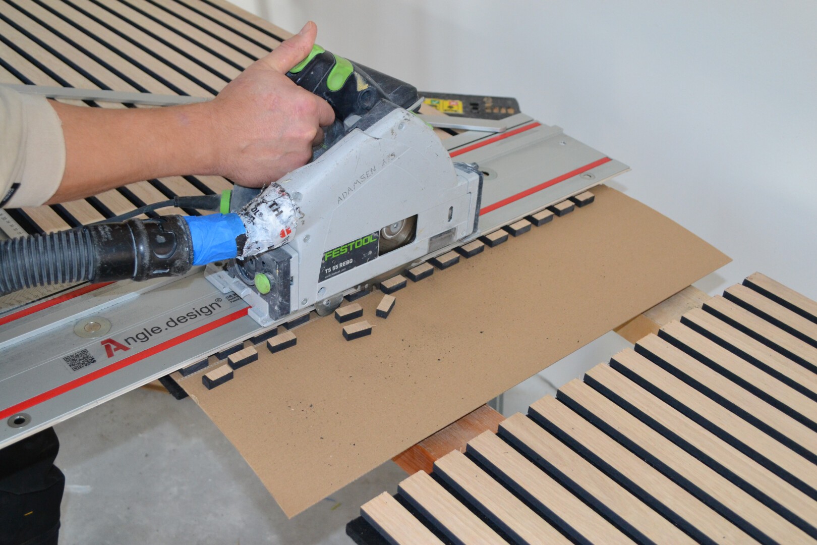 A person is using a circular saw with a guide rail to cut rectangular notches into a sheet of brown material, likely MDF or wood, with slatted wooden pieces nearby.
