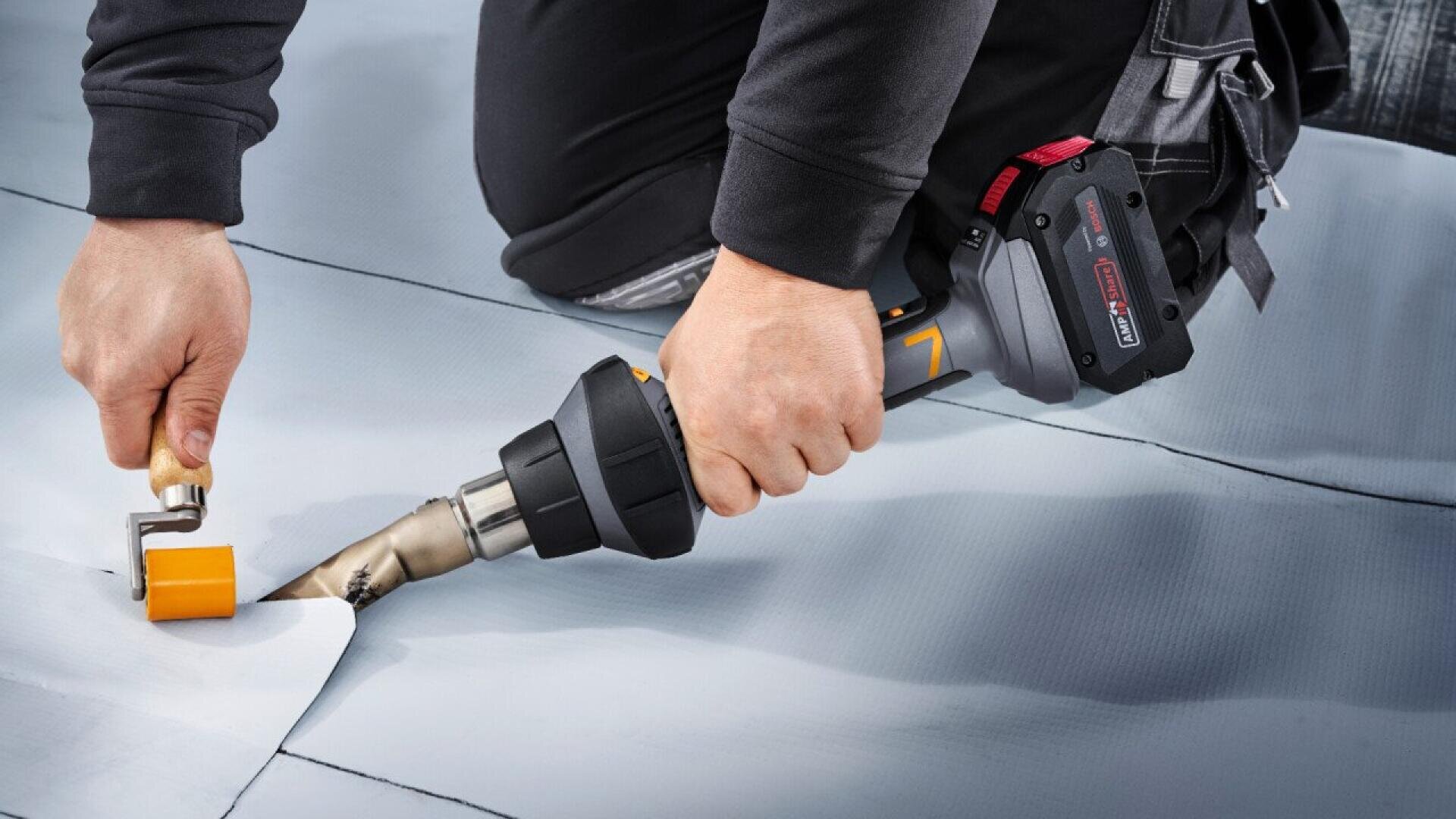 A person uses a heat gun and a roller to weld or seal seams on a gray sheet material, likely performing roofing or flooring installation work.