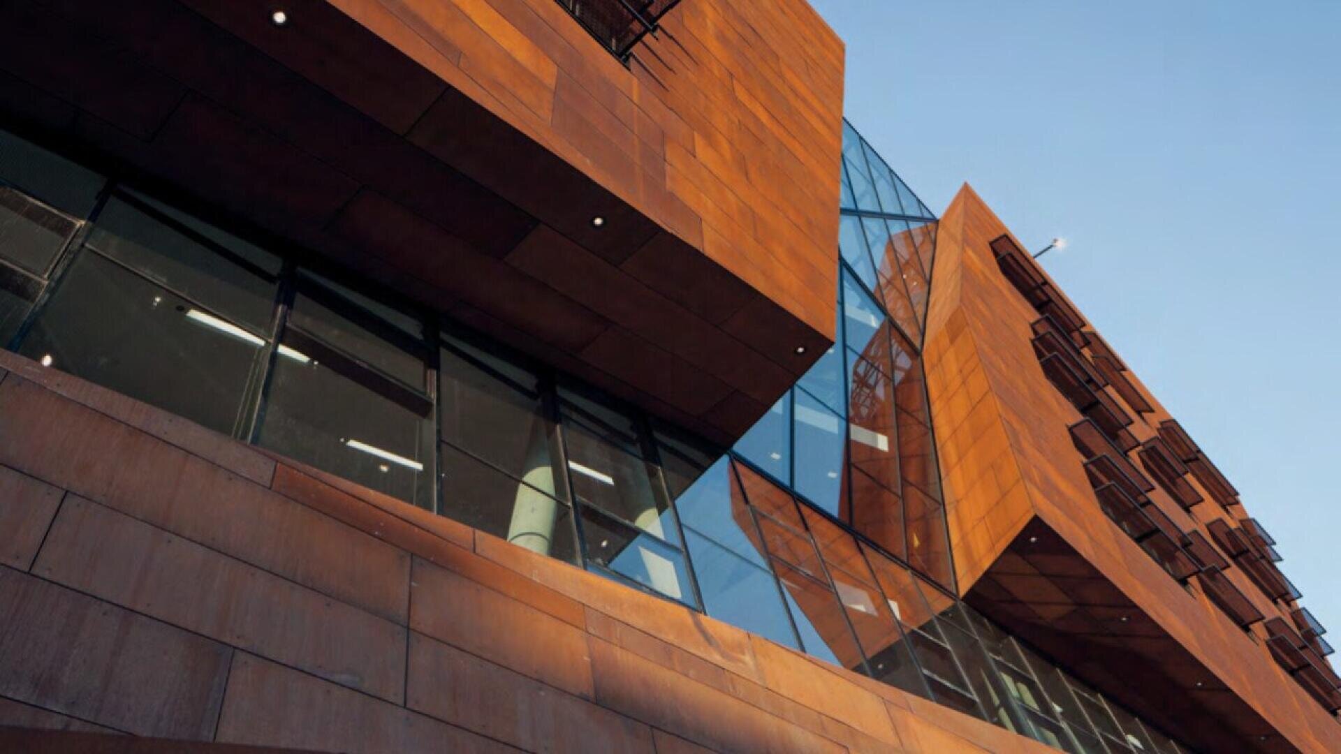A modern building with angular architecture, featuring rust-colored panels and large glass windows, viewed from below against a clear blue sky.