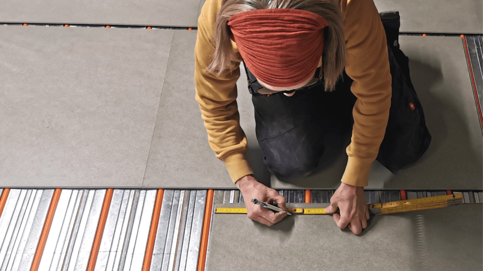 Eine Person mit rotem Stirnband und gelbem Pullover kniet auf dem Boden und misst und markiert eine große Fliese mit Lineal und Bleistift. Die Fliese wird über freiliegende Fußbodenheizungsstreifen verlegt.