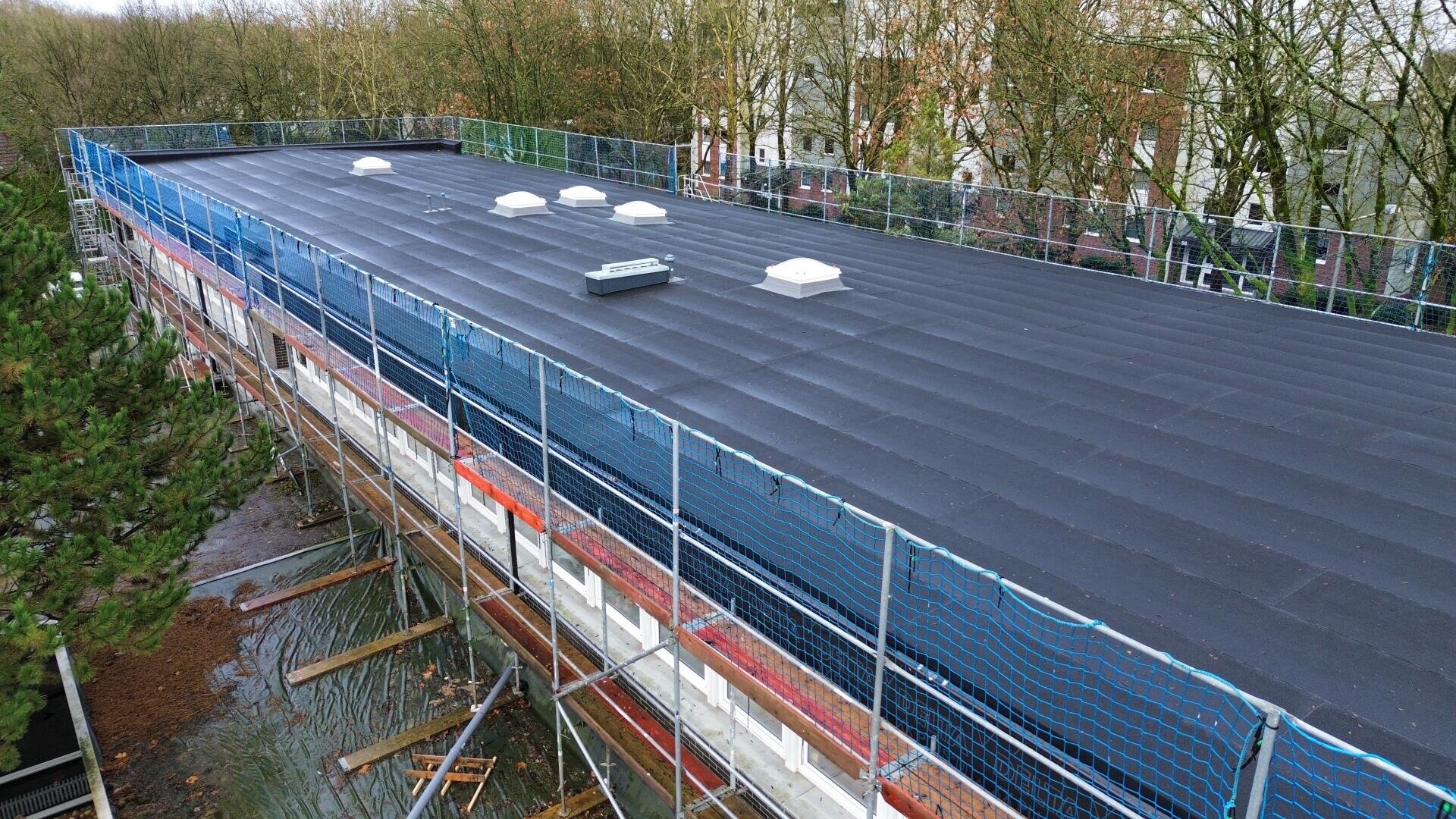 Ein großes, mit schwarzem, wasserdichtem Material bedecktes Flachdach, umgeben von blauen Sicherheitsnetzen und Gerüsten, mit mehreren weißen Dachluken; im Hintergrund sind Bäume und Gebäude zu sehen.