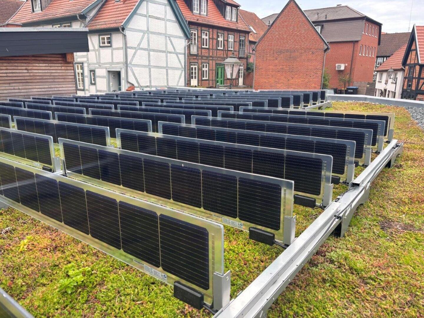 Reihen von Sonnenkollektoren auf einem grünen, moosbewachsenen Dach mit traditionellen europäischen Häusern im Hintergrund.