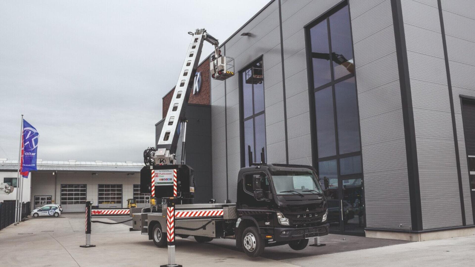 A black truck with an extended articulated boom lift raises a worker in a bucket to a high window on a modern industrial building, with safety stabilizers deployed on both sides.