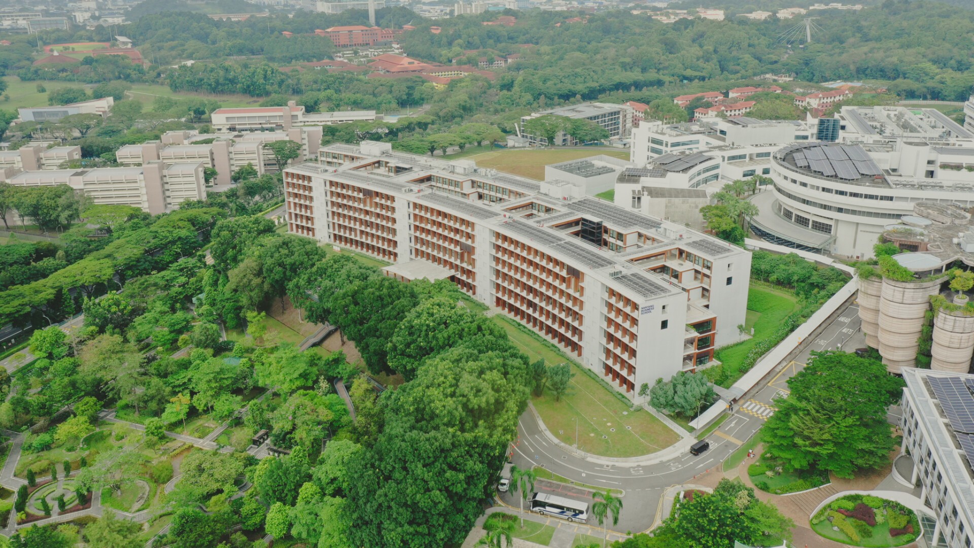 Luftaufnahme eines großen Universitätscampus mit modernen Gebäuden, umgeben von üppigem Grün, kurvigen Straßen und einem Bus in der Nähe des Eingangs. Im Hintergrund sind bewaldete Hügel und weitere akademische Gebäude zu sehen.