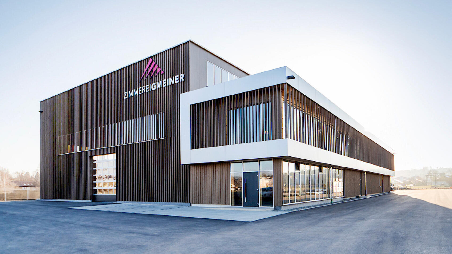Ein modernes Industriegebäude mit Holz- und weißer Verkleidung verfügt über große Fenster und das Schild „ZIMMEREI GEMEINER“ an der Fassade, vor einem klaren Himmel und einer leeren gepflasterten Fläche.