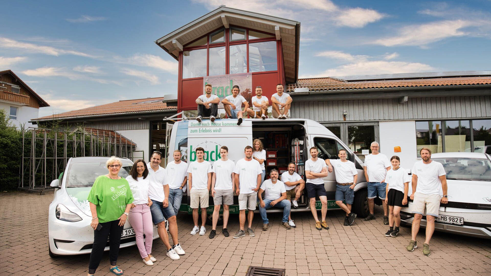 Eine Gruppe von Menschen in weißen Hemden posiert vor einem modernen Gebäude und einem weißen Lieferwagen mit grünem Logo. Einige stehen auf dem Lieferwagen, andere auf dem Boden. Es ist ein sonniger Tag mit blauem Himmel und vereinzelten Wolken.