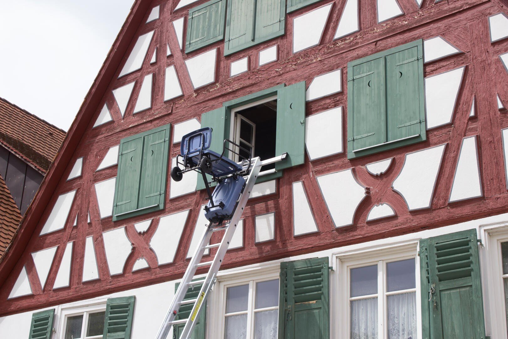 Ein Fahrstuhl wird bis zu einem offenen Fenster im zweiten Stock eines traditionellen Fachwerkhauses mit grünen Fensterläden und dekorativen roten Holzbalken ausgefahren.