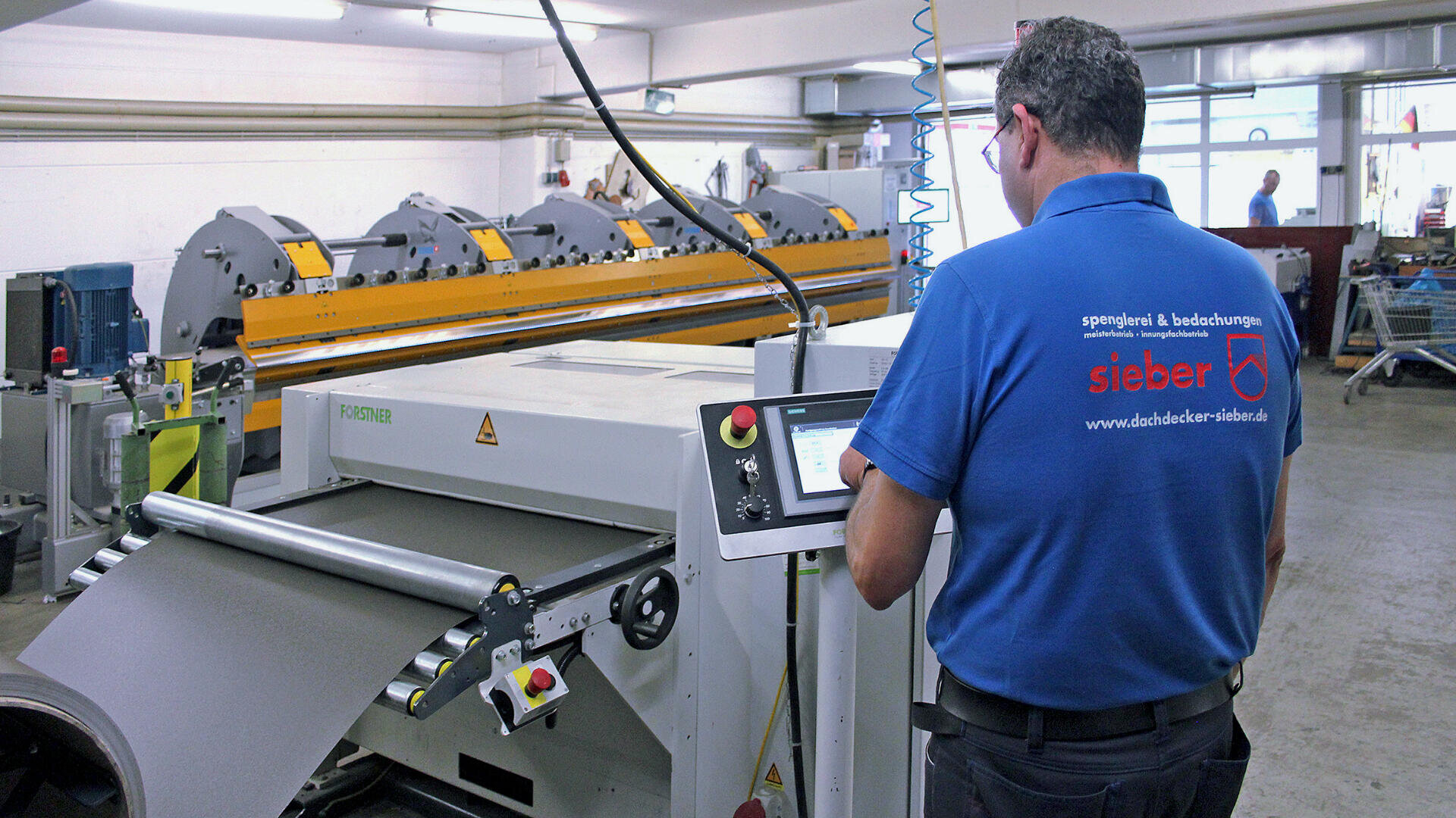 Ein Mann in einem blauen Hemd der Firma Sieber bedient in einer Fabrik ein Touchscreen-Panel. Er steht neben einer großen Maschine zur Blechverarbeitung, im Hintergrund sind Industrieanlagen zu sehen.