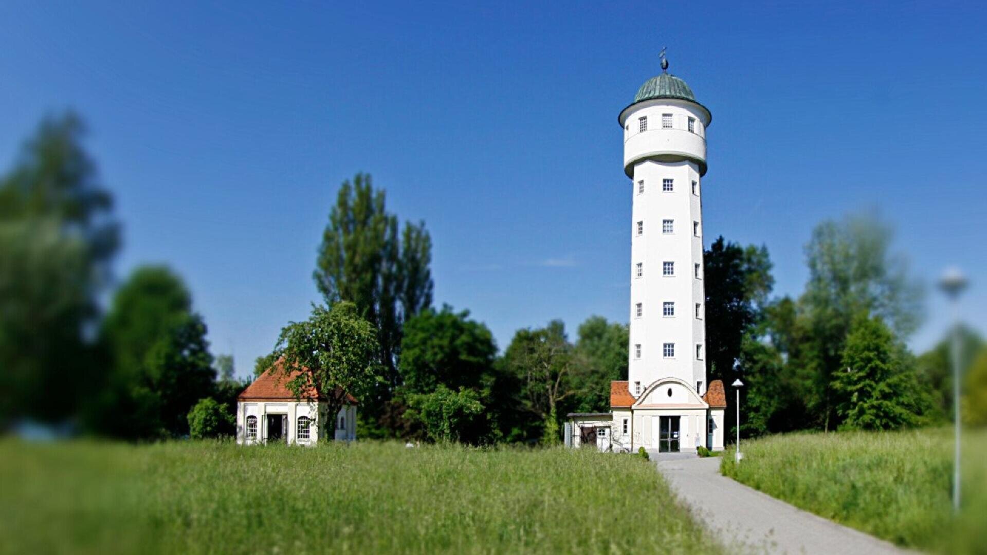 A tall, white cylindrical tower with a green domed roof stands next to a small building with a red roof, surrounded by grass, trees, and a clear blue sky. A pathway leads to the tower entrance.