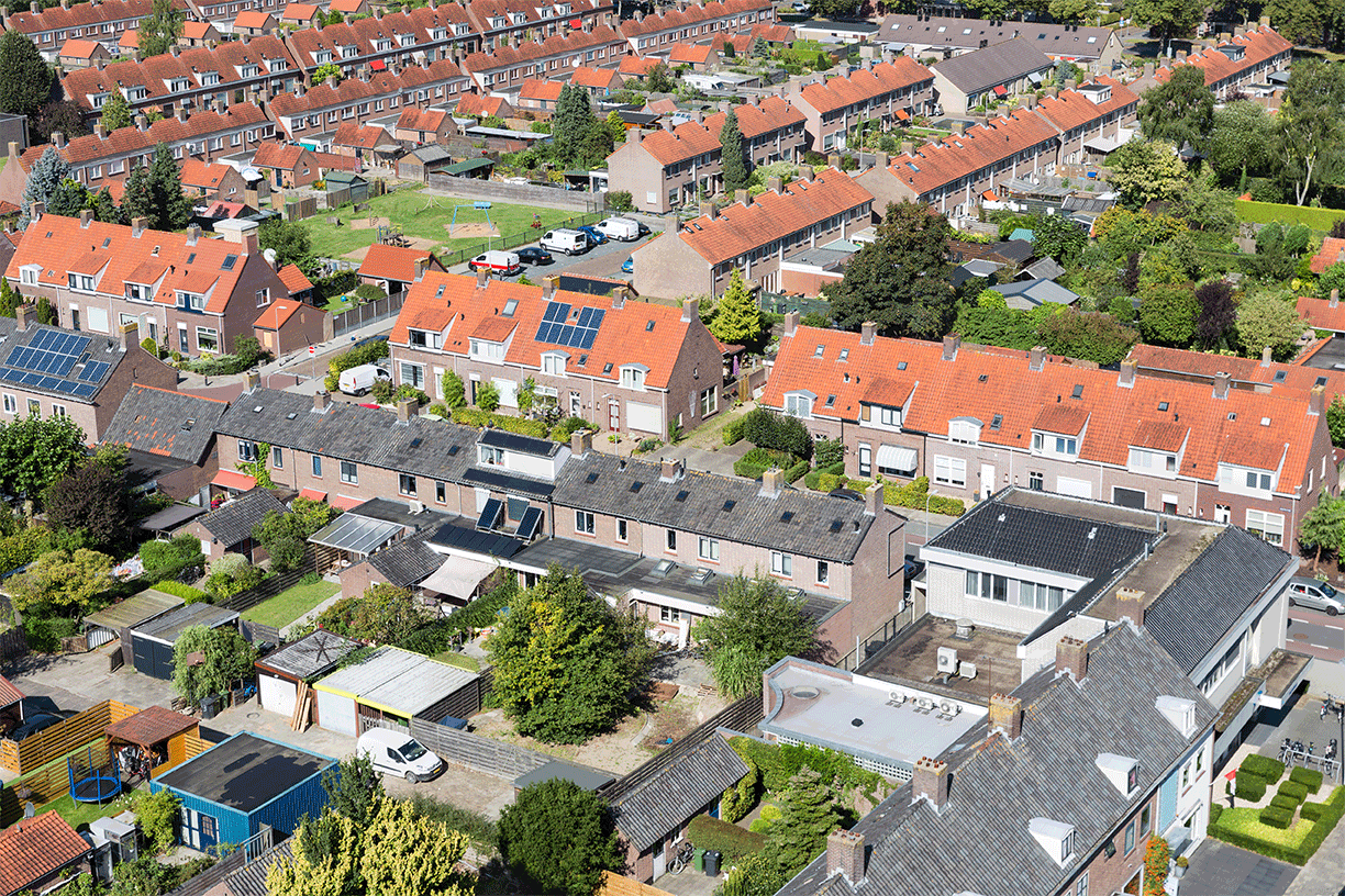Luftaufnahme eines Wohnviertels mit Häuserreihen mit roten Ziegeldächern, grünen Gärten, Solarmodulen und von Bäumen gesäumten Straßen an einem sonnigen Tag.
