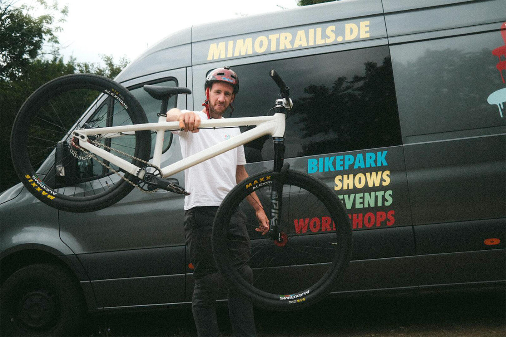 Ein Mann mit Helm hält ein weißes Mountainbike am Rahmen hoch und steht vor einem dunklen Lieferwagen mit der Aufschrift MIMOTRAILS.DE und bunten Wörtern wie BIKEPARK, SHOWS, EVENTS und WORKSHOPS auf der Seite.