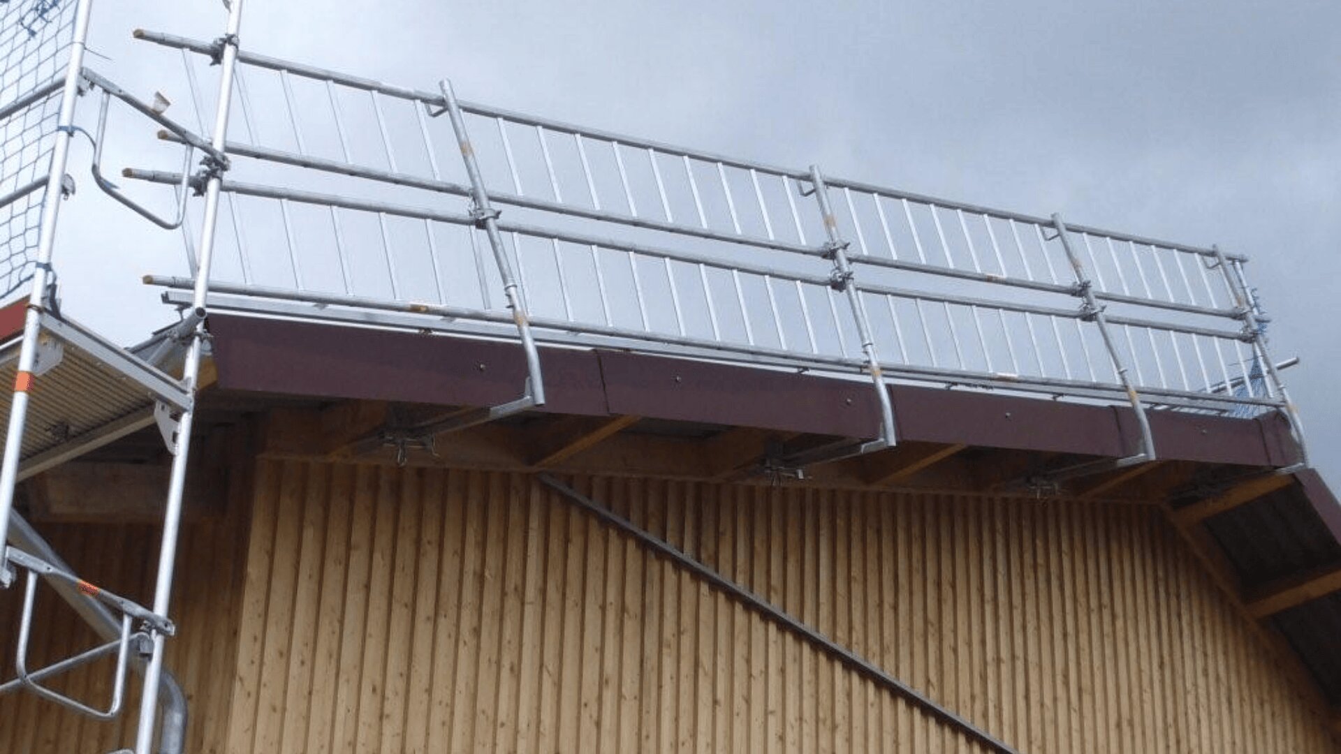 Entlang der Kante eines schrägen Holzdachs ist ein Metallgerüst mit Sicherheitsgeländern aufgestellt, im Hintergrund ist ein bewölkter Himmel zu sehen.