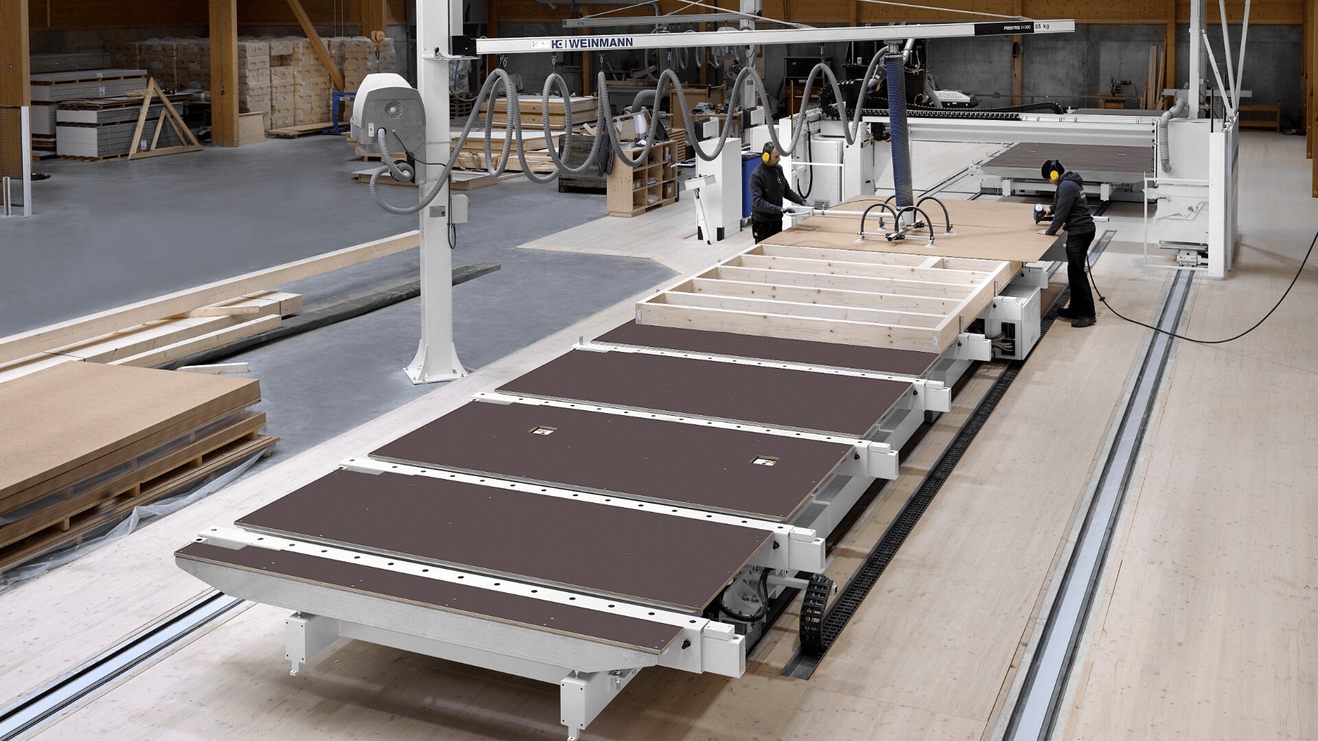 Zwei Arbeiter bedienen eine große automatisierte Maschine, die Holzwandpaneele in einer geräumigen, modernen Fabrik zusammenbaut. In der Nähe sind Holzmaterialien gestapelt.