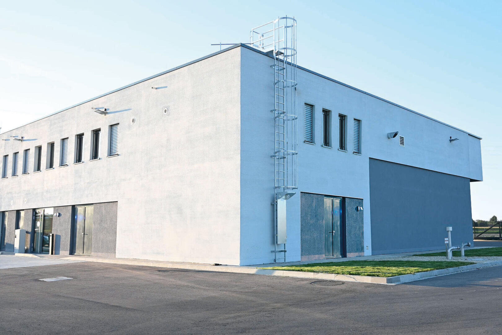 Ein modernes, minimalistisches zweistöckiges Gebäude mit grau-weißen Außenwänden, mehreren Fenstern und einer Metallleiter an der Seite, gelegen an einer gepflasterten Straße und einem kleinen Grasfleck unter klarem Himmel.