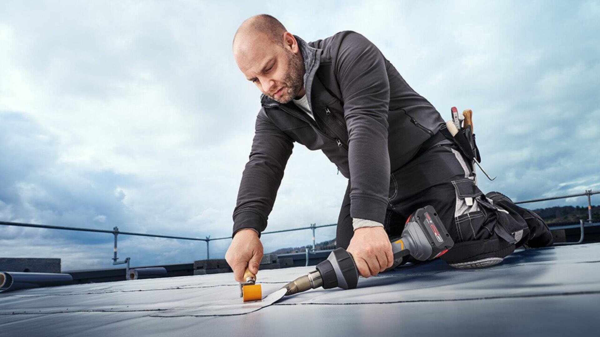 A person kneels on a rooftop, using a handheld heat gun and roller to seal roofing material. They wear work clothes with tools in a belt, and the sky is cloudy in the background.