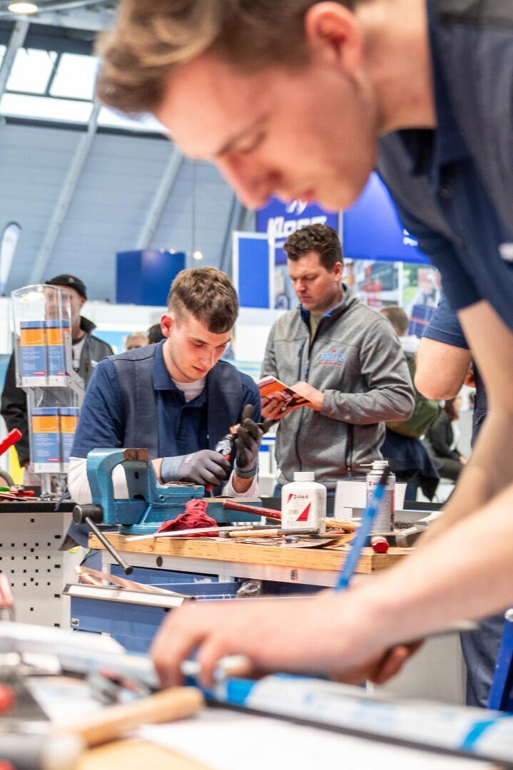 Eine Gruppe junger Männer arbeitet an technischen Projekten an einem Arbeitsplatz. Einer konzentriert sich auf die Werkzeuge in seinen Händen, ein anderer schreibt im Vordergrund, und andere beobachten im Hintergrund, was eine Werkstatt oder eine Messe zu sein scheint.
