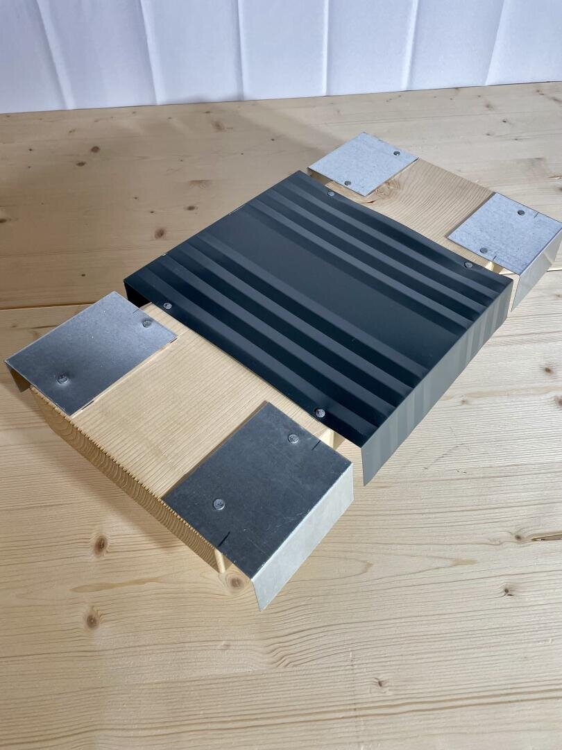 Ein Holztisch zeigt eine geometrische Struktur aus hellem Holz, Metallplatten und einer zentralen schwarzen Platte mit erhabenen parallelen Streifen. Die Struktur zeichnet sich durch scharfe Winkel und sichtbare Schrauben aus.