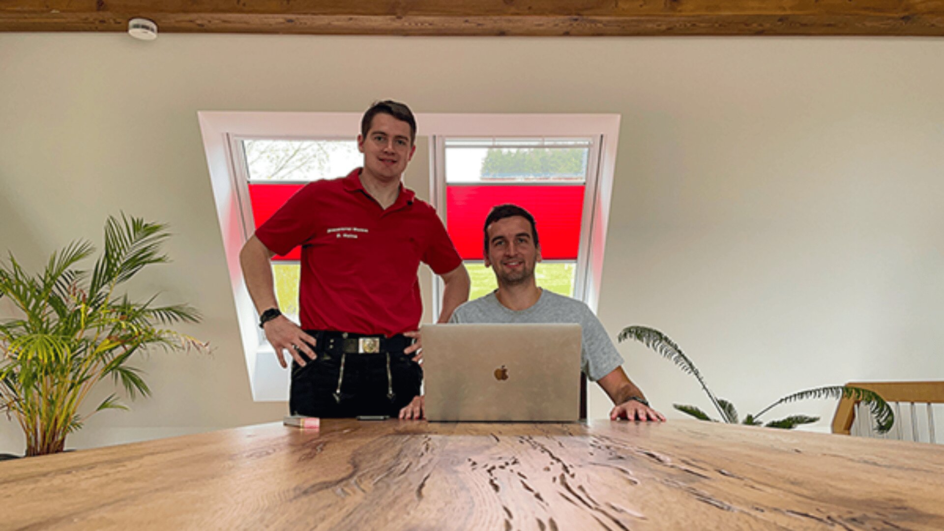 Zwei Männer sitzen an einem Holztisch, einer steht, der andere sitzt mit einem Laptop. Zu beiden Seiten stehen grüne Pflanzen, im Hintergrund ein Fenster mit roten Jalousien. Die Atmosphäre ist entspannt und hell.