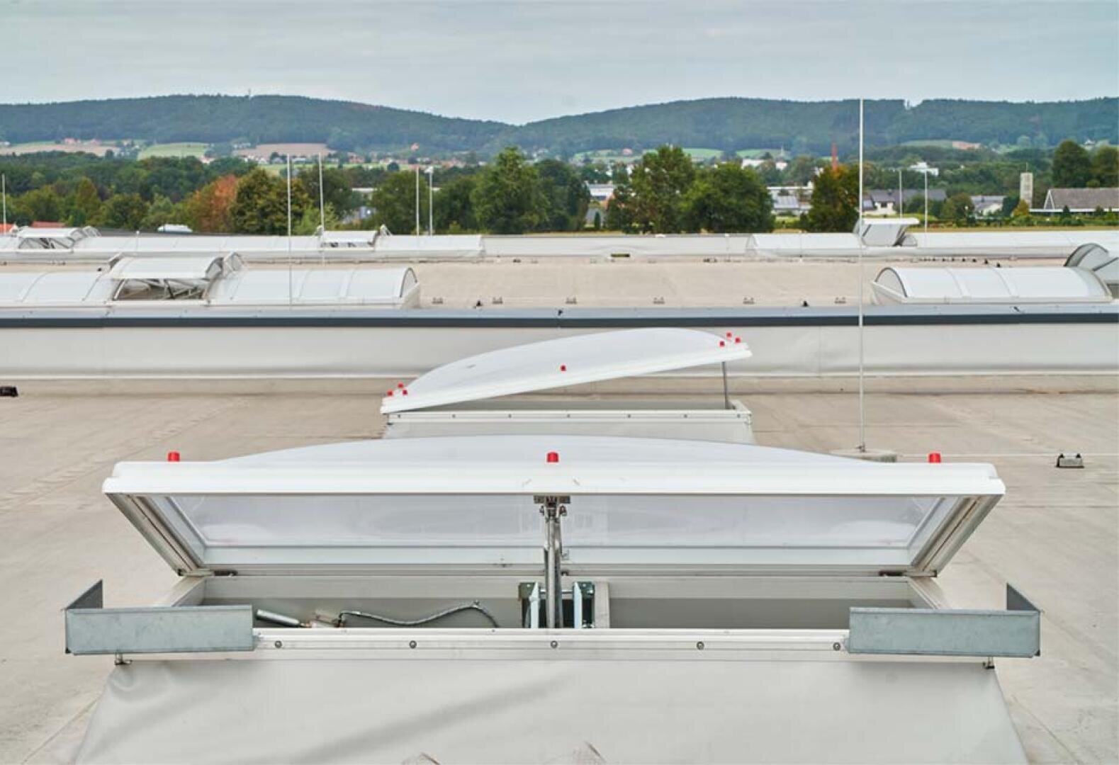 A flat industrial roof with several rectangular white chimneys, some open, some closed, surrounded by trees and distant hills under a cloudy sky.