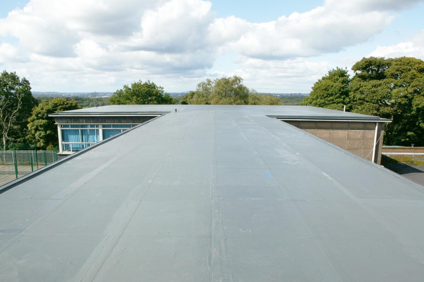 Blick auf ein flaches, graues Dach, das sich in die Ferne erstreckt, umgeben von grünen Bäumen und einem teilweise bewölkten Himmel, wobei links ein Teil eines modernen Gebäudes zu sehen ist.