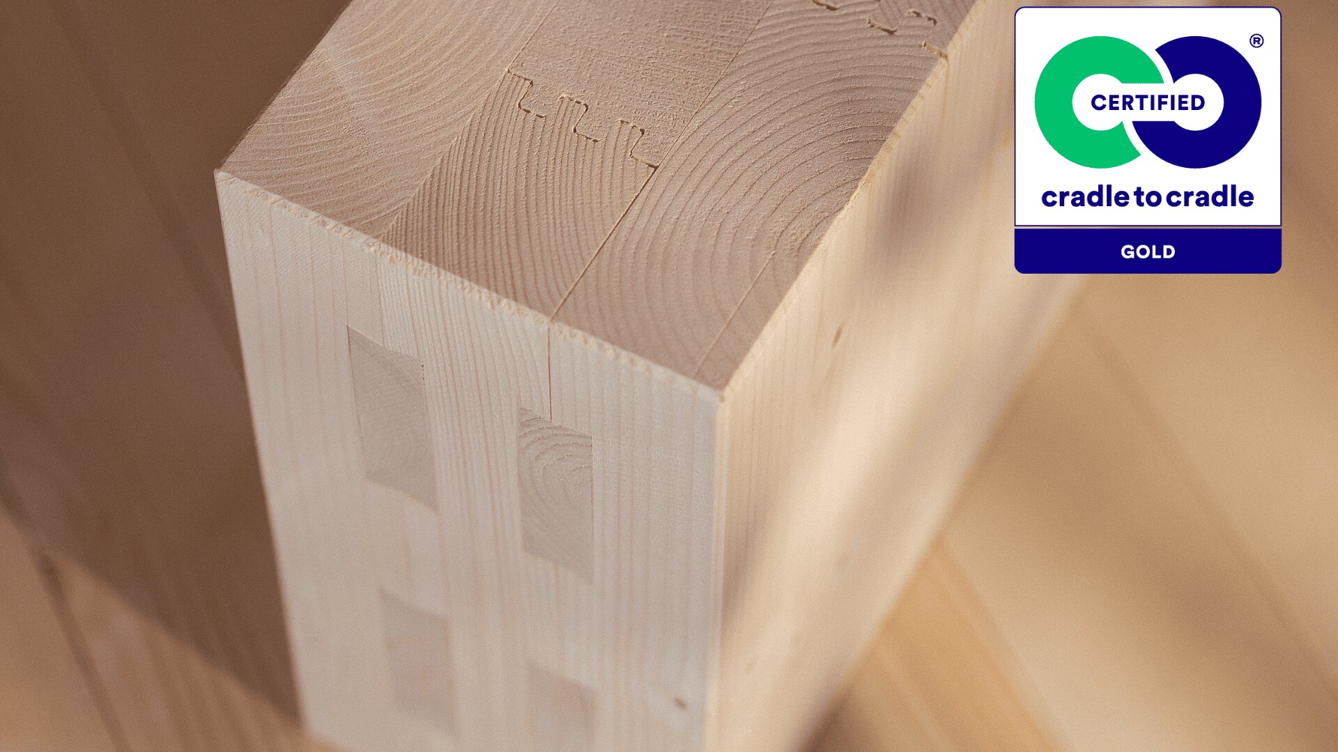 Nahaufnahme eines glatten, hellen Holzblocks mit ineinandergreifenden Verbindungen und dem „Cradle to Cradle Certified Gold“-Logo in der oberen rechten Ecke. Der Hintergrund zeigt weitere helle Holzoberflächen.
