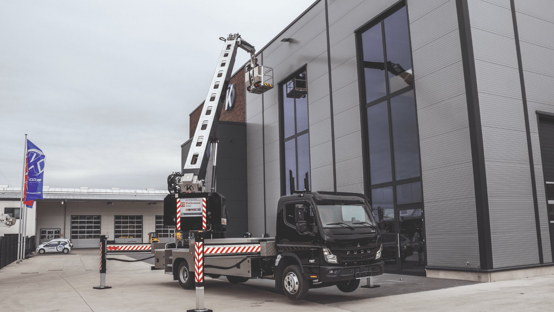 Ein schwarzer LKW mit montiertem Kran fährt Arbeitern eine Plattform aus, die die Außenseite eines modernen Industriegebäudes mit großen Fenstern inspizieren oder reparieren.