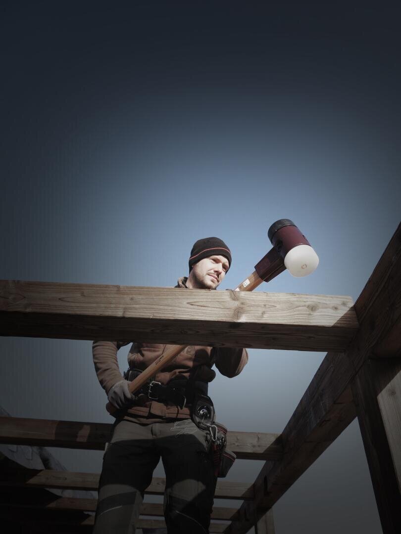 Ein Bauarbeiter mit Mütze und Handschuhen benutzt unter klarem Himmel einen großen Gummihammer, um Holzbalken an einem Gebäuderahmen zu richten.