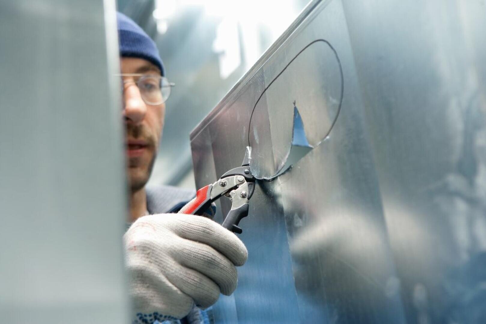 A person wearing gloves and a beanie uses metal snips to cut an oval shape in a sheet of metal. The focus is on the hands and tool, with the face blurred in the background.