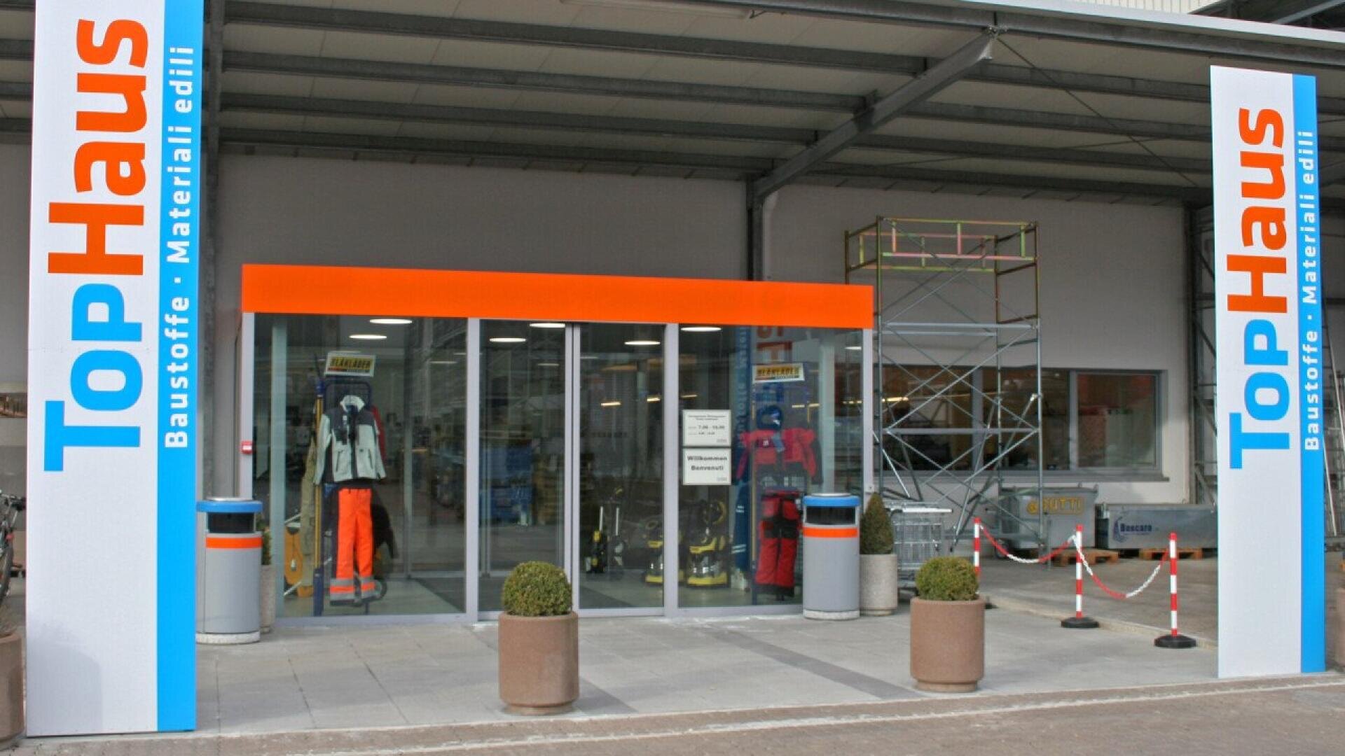 Entrance of a TopHaus hardware store with glass doors, safety gear display, potted plants, and scaffolding equipment visible. Store signs in German and Italian are displayed on tall columns by the entrance.